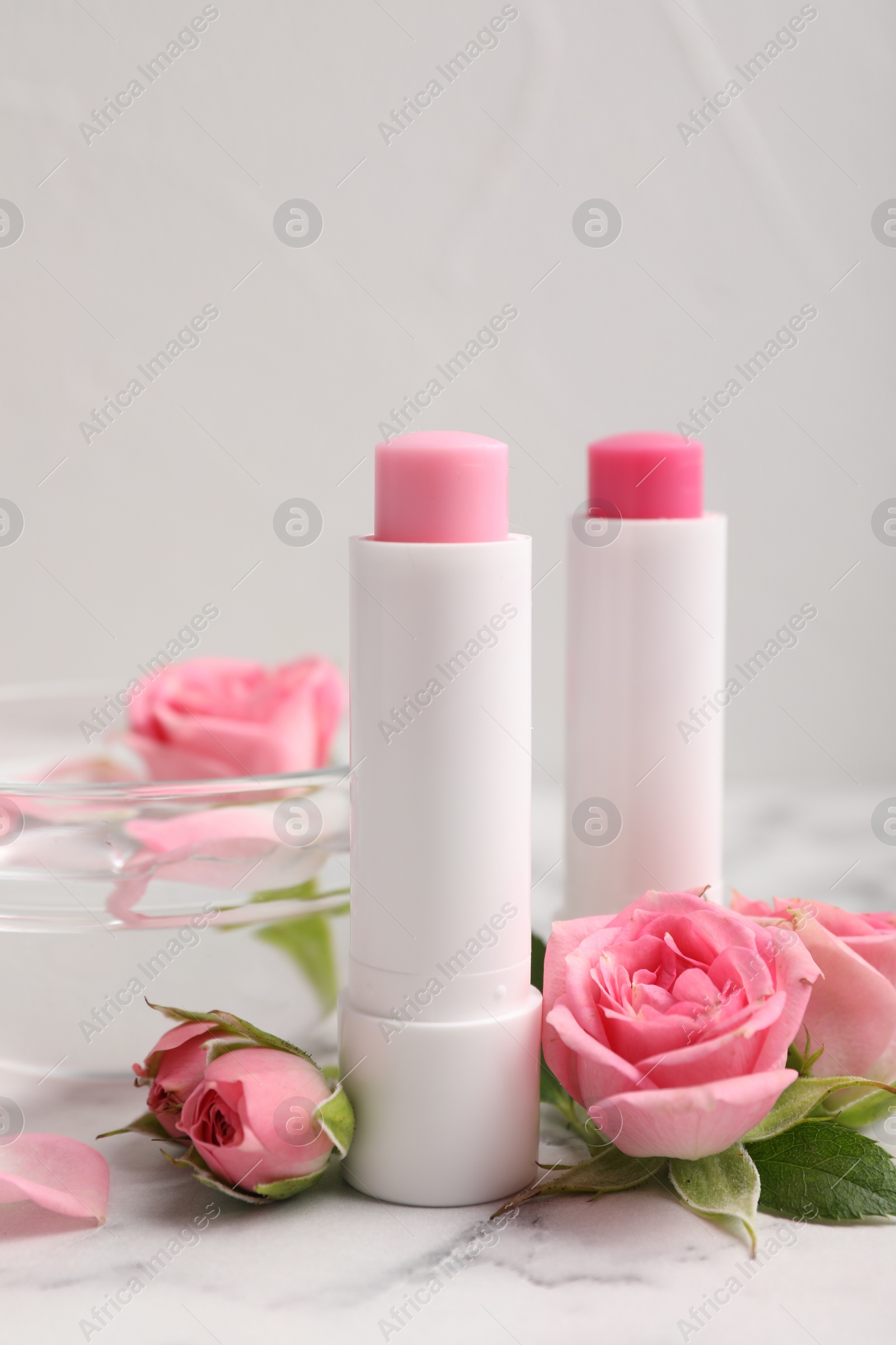 Photo of Lip balms, bowl with water and roses on white marble table