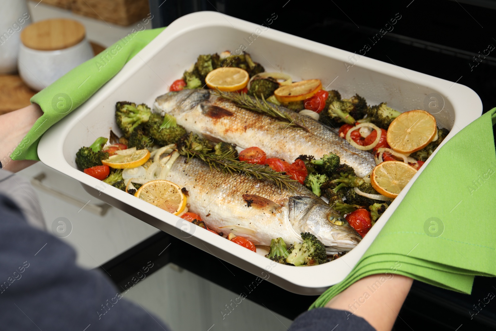 Photo of Woman taking baking dish with delicious fish and vegetables from oven in kitchen, closeup