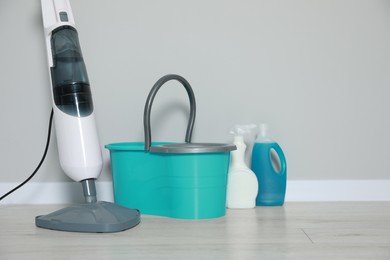 Modern steam mop and bucket with different cleaning supplies on floor near grey wall