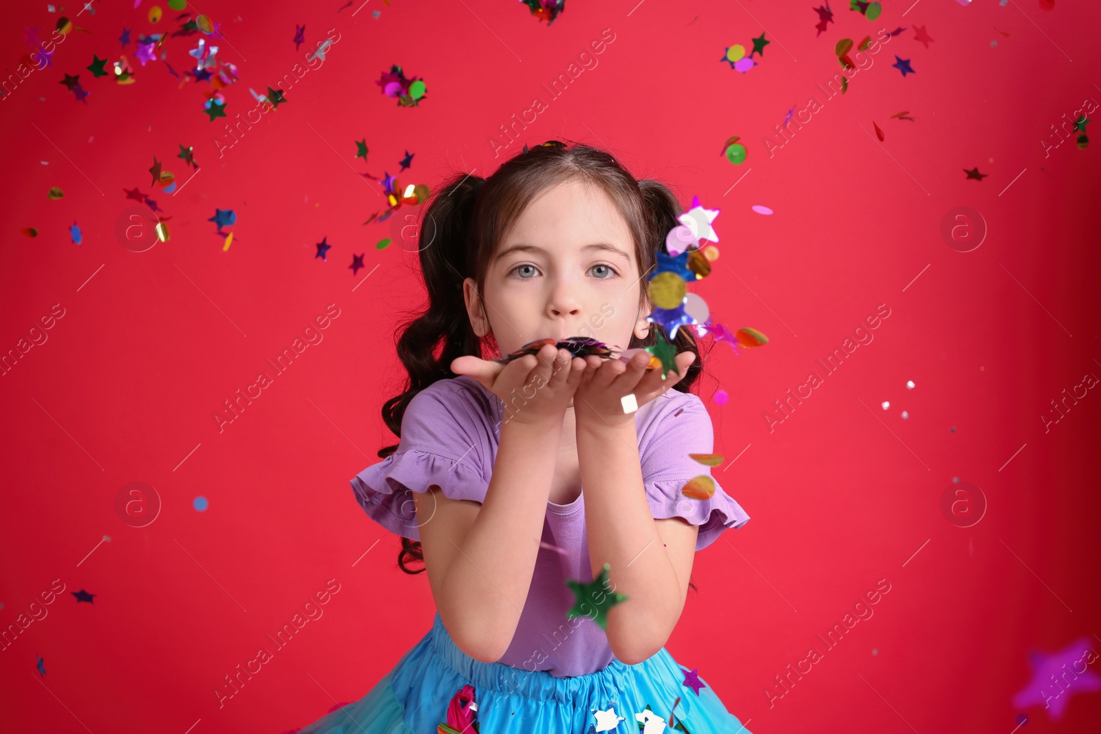 Photo of Adorable little girl blowing confetti on red background
