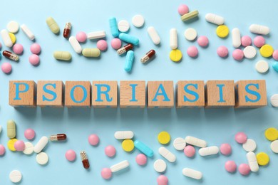 Word Psoriasis made of wooden cubes with letters and different pills on light blue background, flat lay