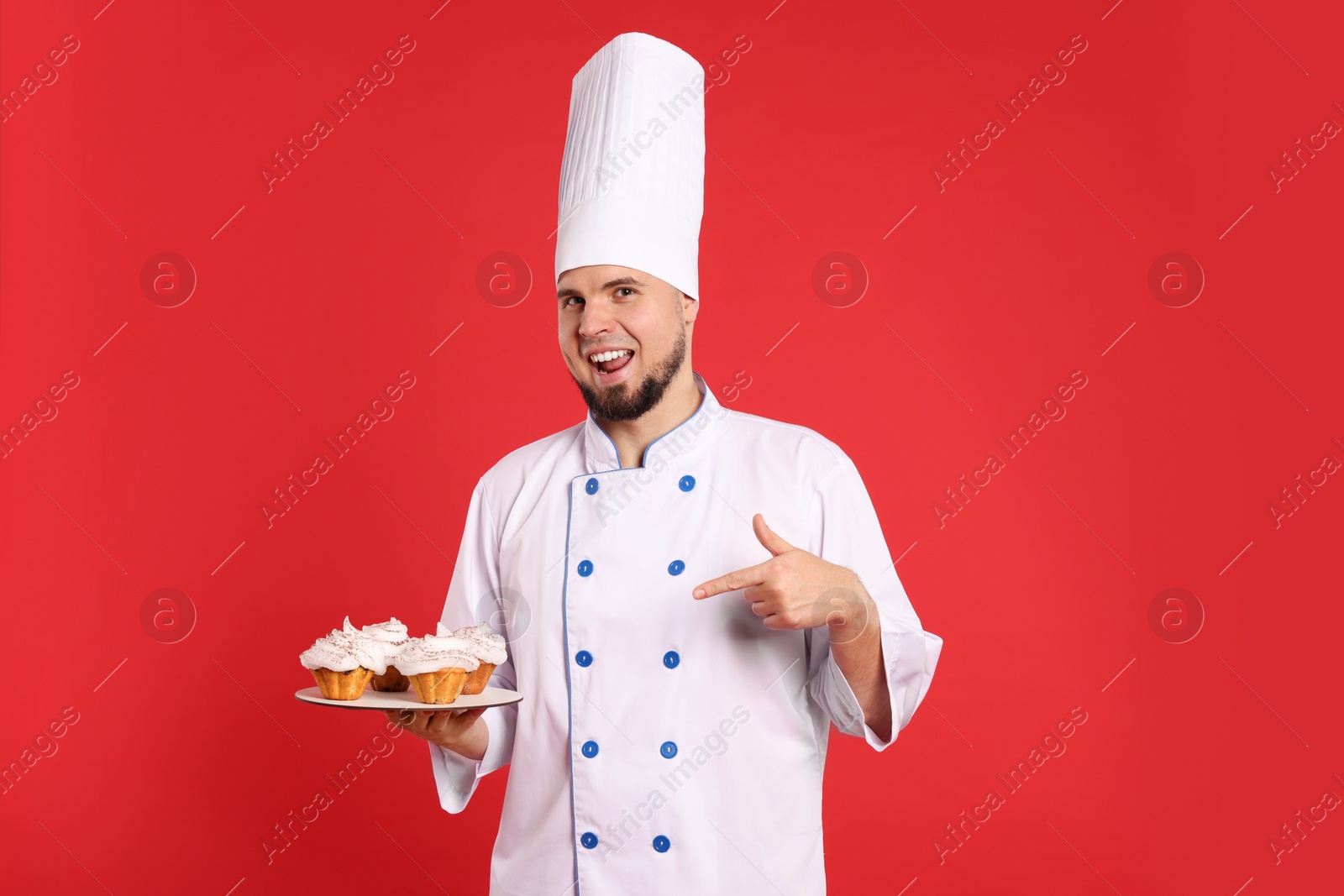 Photo of Happy professional confectioner in uniform pointing at delicious cupcakes on red background