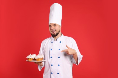 Photo of Happy professional confectioner in uniform pointing at delicious cupcakes on red background