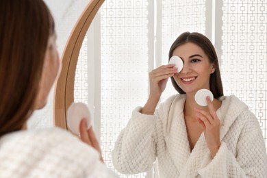 Photo of Young woman with cotton pads cleaning her face near mirror in bathroom