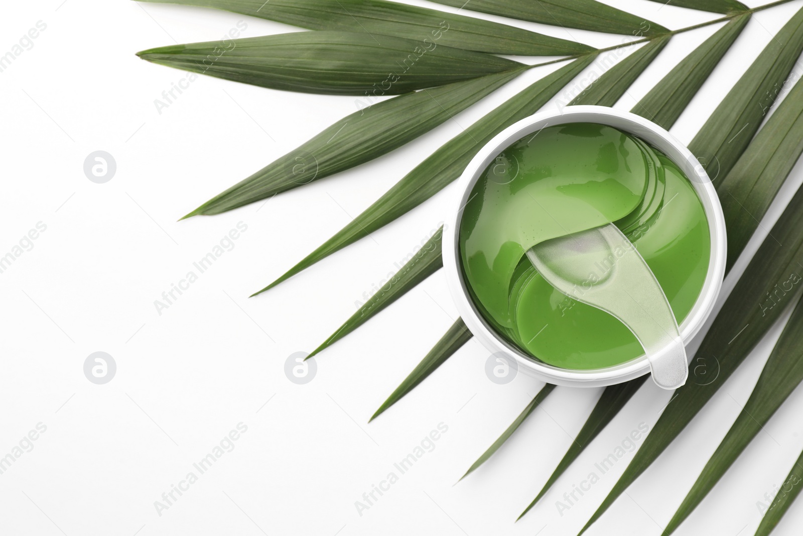 Photo of Jar of under eye patches with spoon and palm leaf on white background, top view with space for text. Cosmetic product