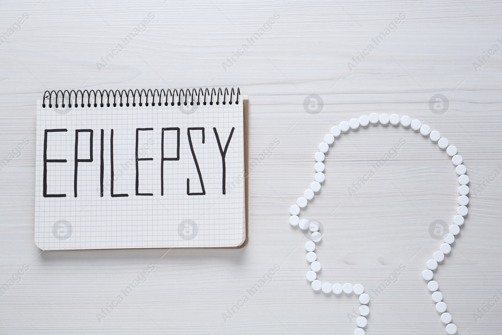 Photo of Human head silhouette made of pills and notebook with word Epilepsy on white wooden table, flat lay