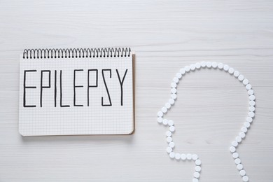 Photo of Human head silhouette made of pills and notebook with word Epilepsy on white wooden table, flat lay