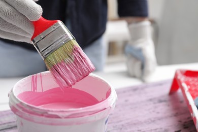 Woman dipping brush into bucket of pink paint indoors, closeup. Space for text