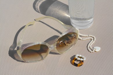 Photo of Stylish sunglasses, glass of water and jewelry on grey surface, closeup