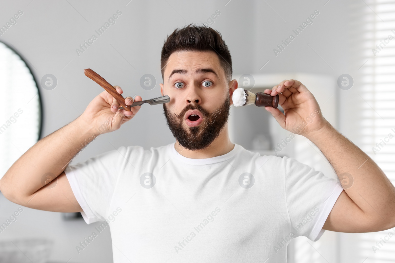 Photo of Handsome young man holding blade and shaving brush with foam in bathroom