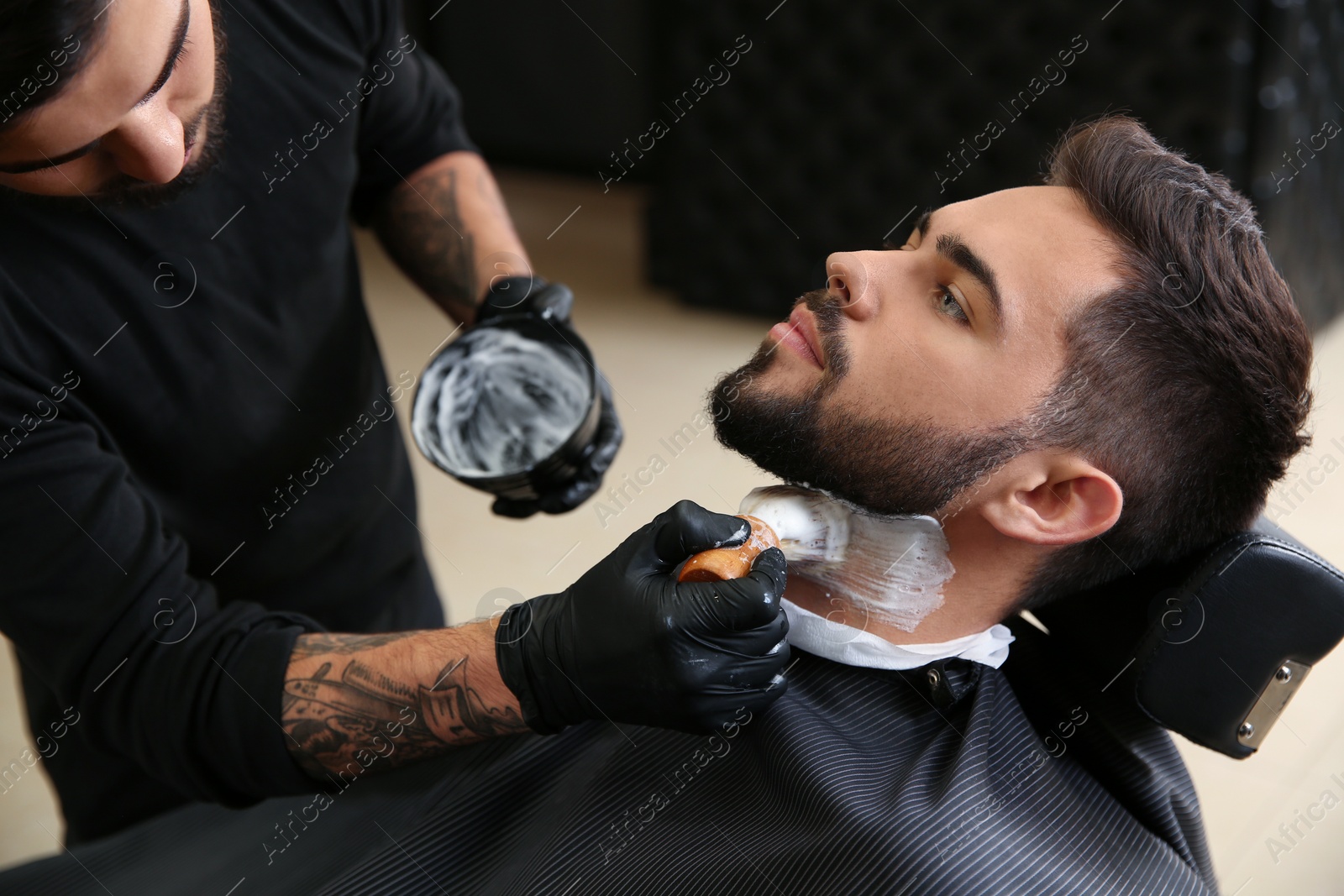 Photo of Professional hairdresser applying shaving foam onto client's skin in barbershop