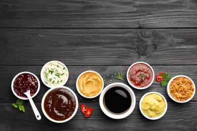 Different tasty sauces in bowls, parsley, chili pepper and rosemary on black wooden table, flat lay. Space for text