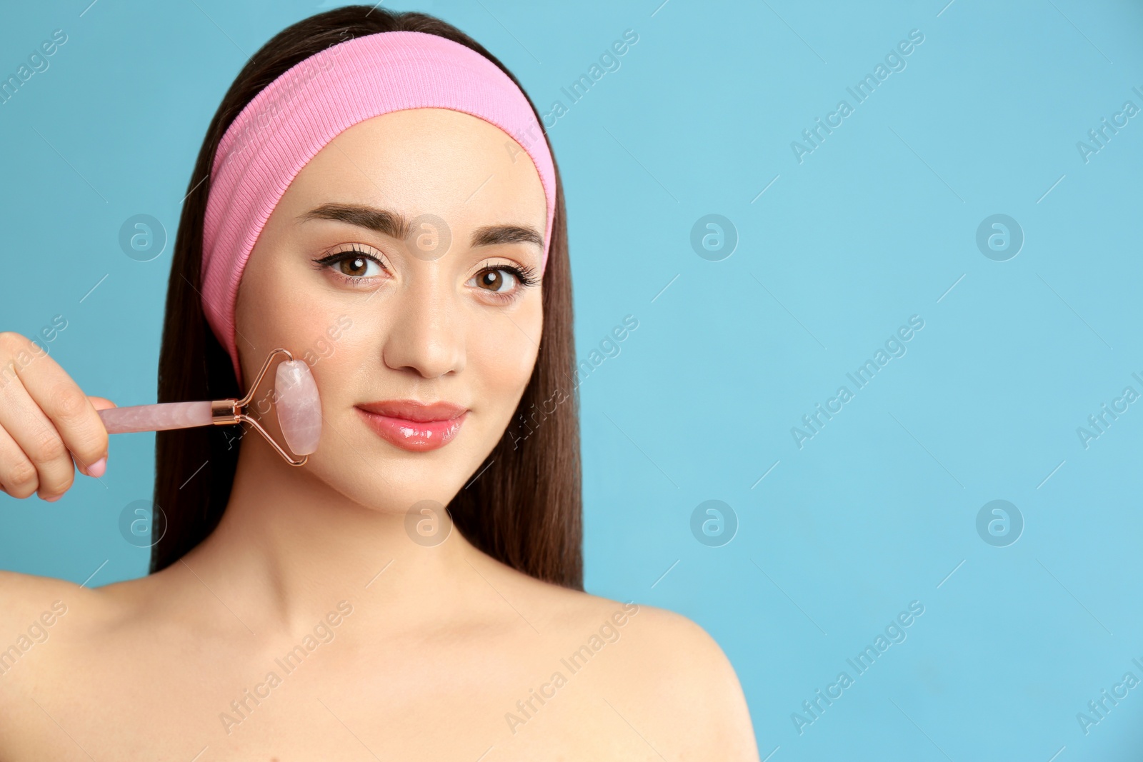 Photo of Woman using natural pink quartz face roller on light blue background, space for text