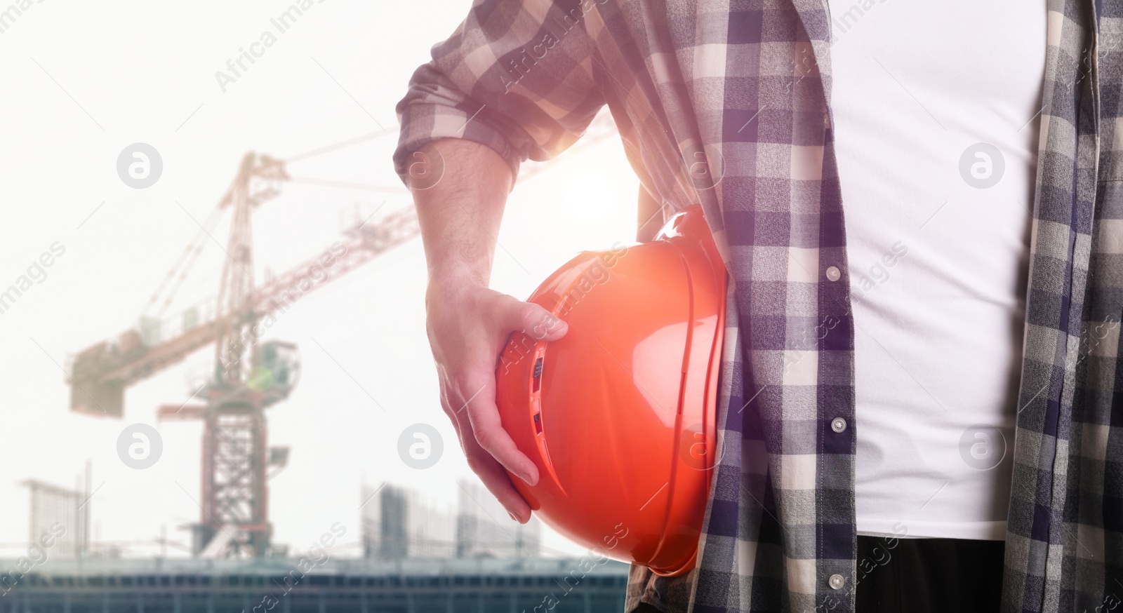 Image of Man holding orange hard hat at construction site with unfinished building, closeup. Space for text 