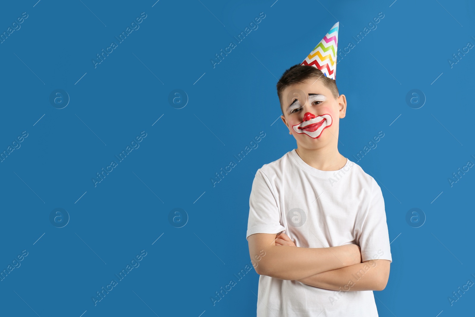 Photo of Preteen boy with clown makeup and party hat on blue background, space for text. April fool's day