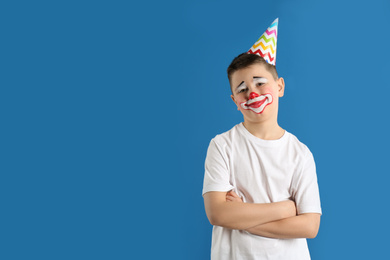 Photo of Preteen boy with clown makeup and party hat on blue background, space for text. April fool's day