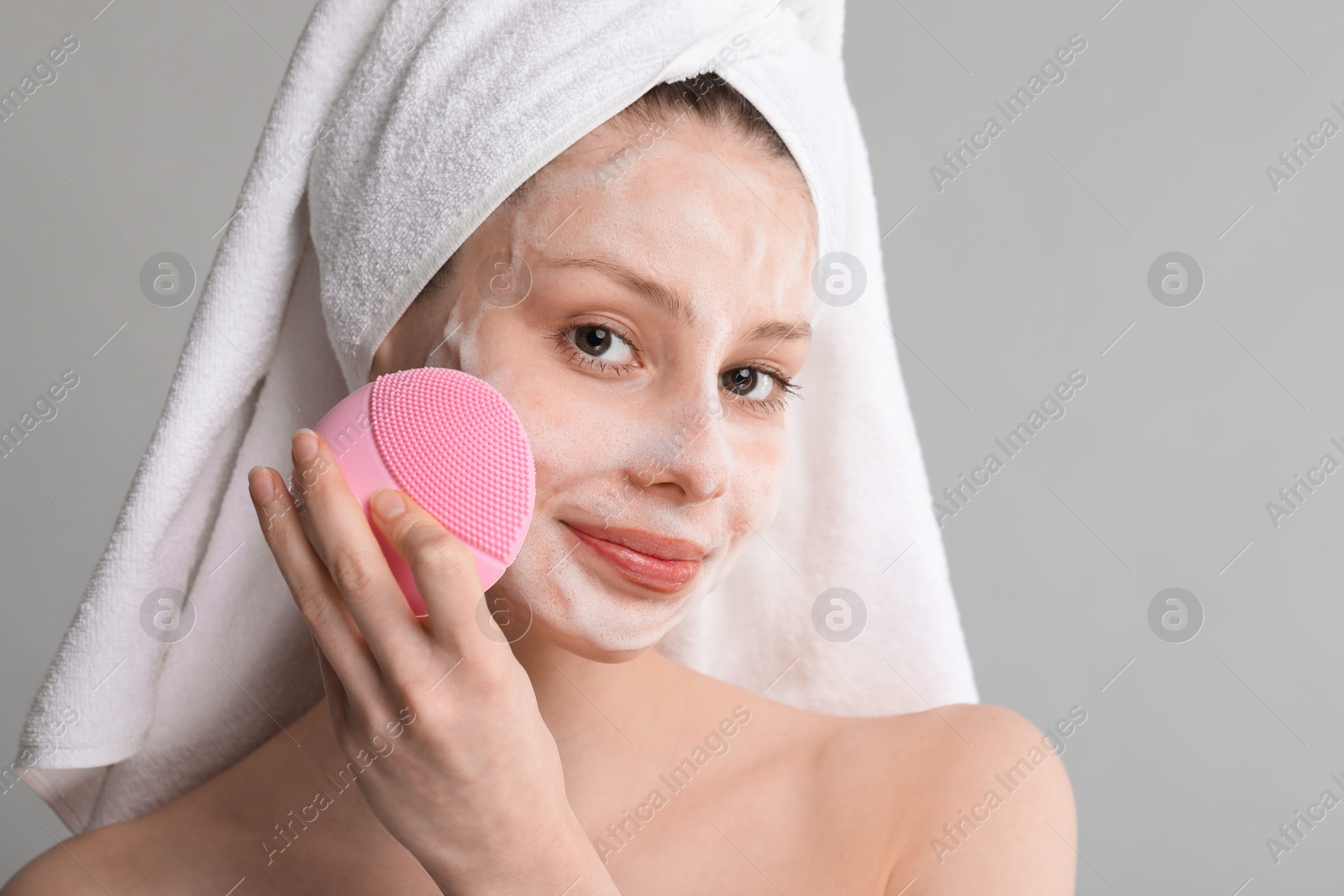 Photo of Young woman washing face with brush and cleansing foam on grey background