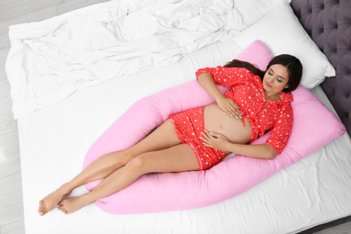 Beautiful pregnant woman lying with maternity pillow on bed at home, top view