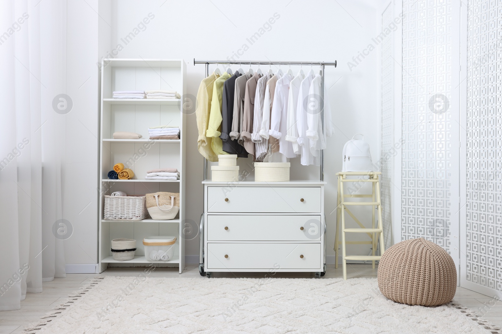 Photo of Wardrobe organization. Rack with different stylish clothes, chest of drawers and folding ladder near white wall indoors