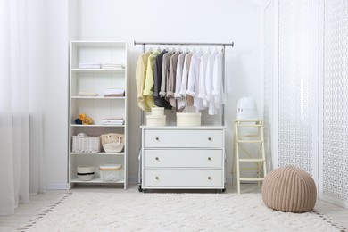 Photo of Wardrobe organization. Rack with different stylish clothes, chest of drawers and folding ladder near white wall indoors