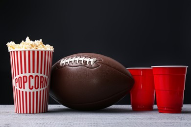 Photo of American football ball with popcorn and cups on grey wooden table