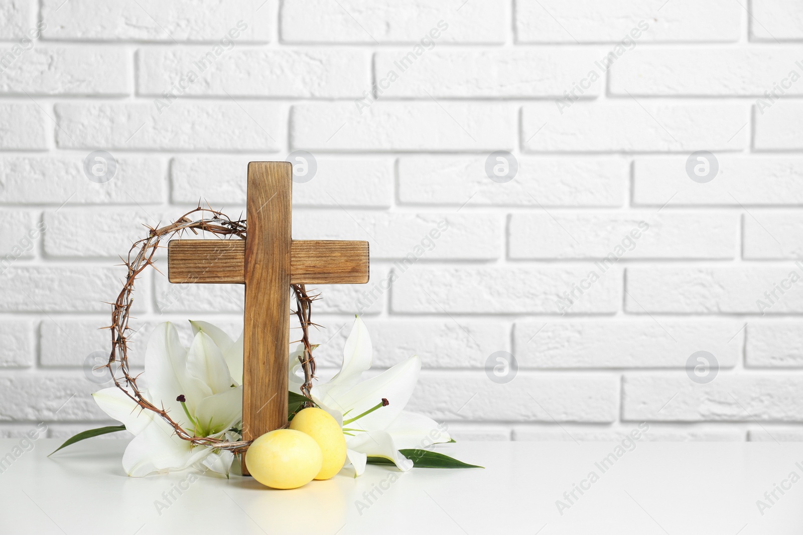 Photo of Wooden cross, crown of thorns, Easter eggs and blossom lilies on table against brick wall, space for text