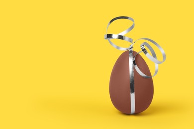 Delicious chocolate egg with silver ribbon on yellow background, closeup. Space for text