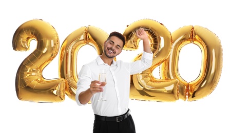 Happy young man with glass of champagne near golden 2020 balloons on white background. New Year celebration