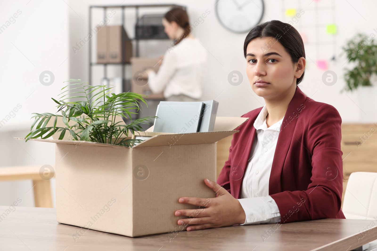 Photo of Unemployment problem. Frustrated woman with box of personal belongings at table in office