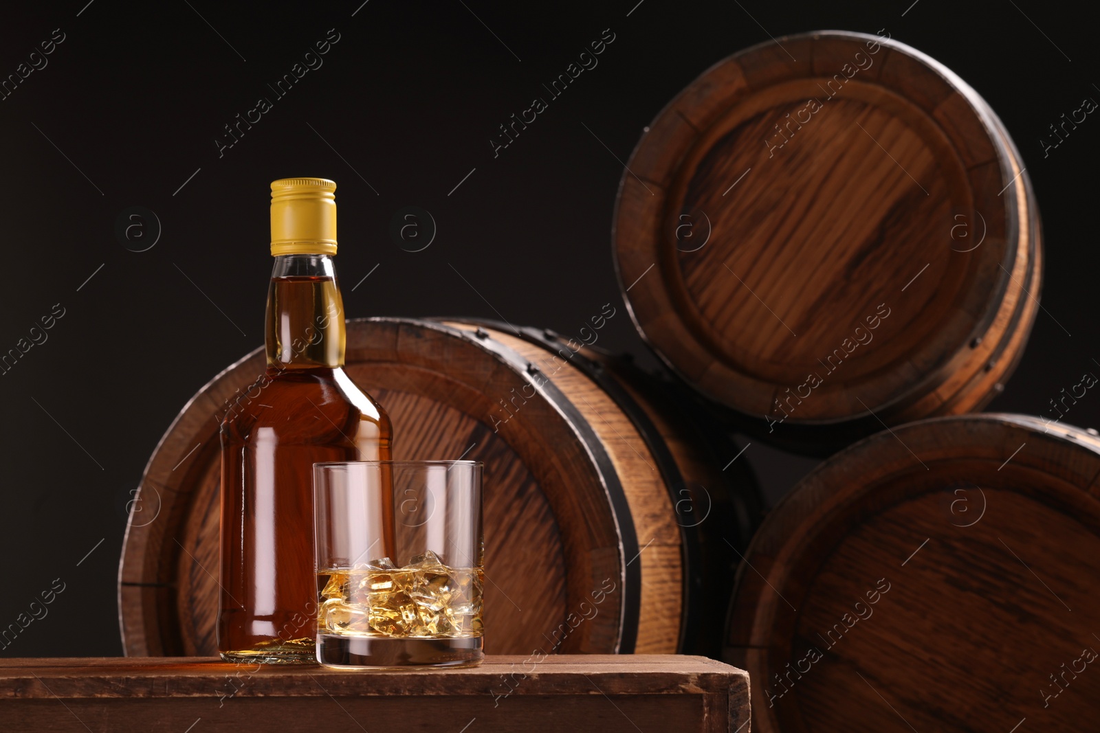 Photo of Whiskey with ice cubes in glass and bottle on wooden table near barrels against black background, space for text