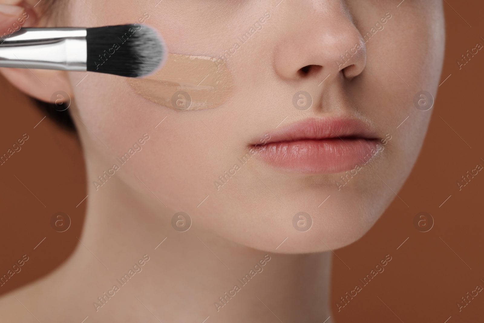 Photo of Teenage girl applying foundation on face with brush against brown background, closeup