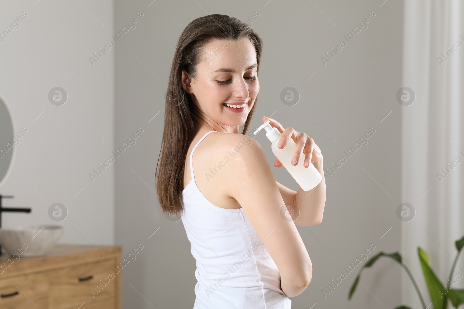 Photo of Happy woman applying body oil onto shoulder in bathroom