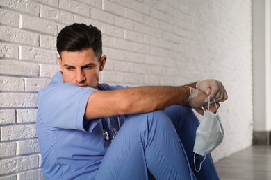 Photo of Sad doctor with protective mask sitting near white brick wall indoors. Stress of health care workers during coronavirus pandemic