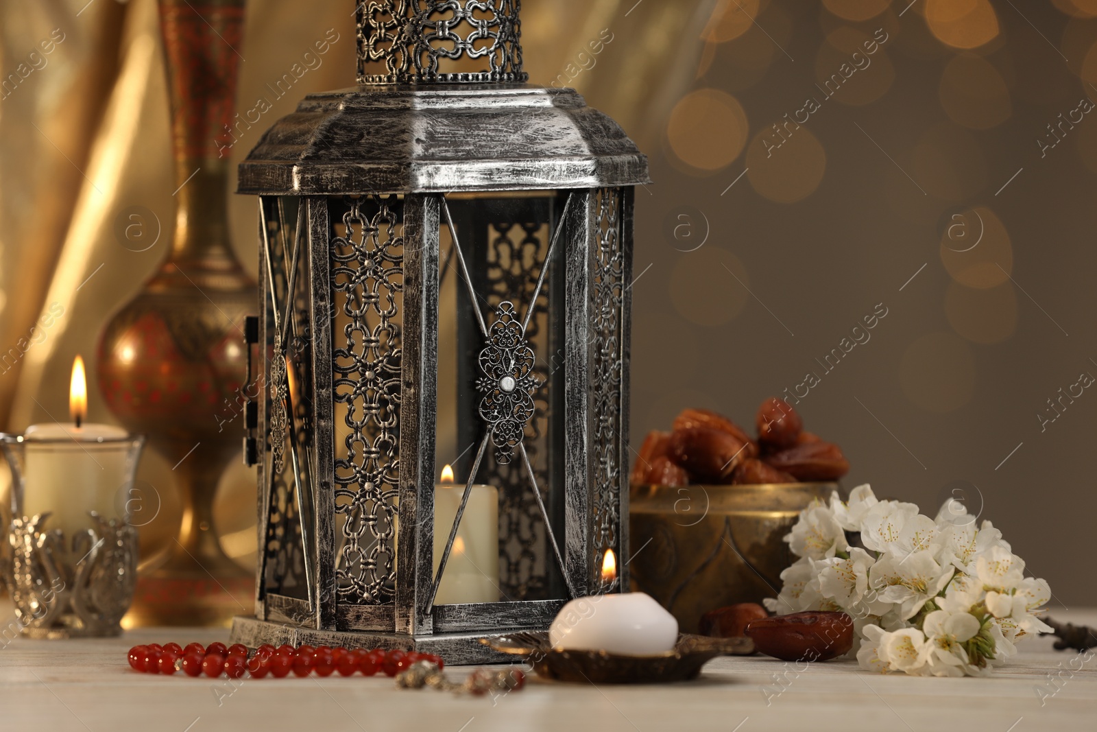 Photo of Arabic lantern, misbaha, candles, dates and flowers on table against blurred lights