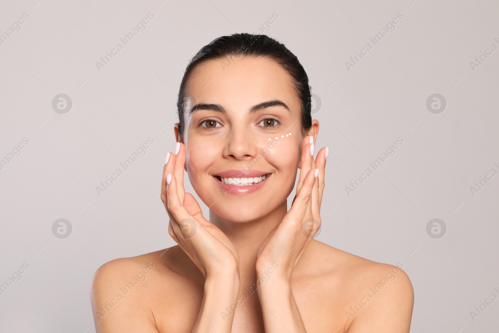 Photo of Beautiful young woman with cream on skin under eye against light grey background