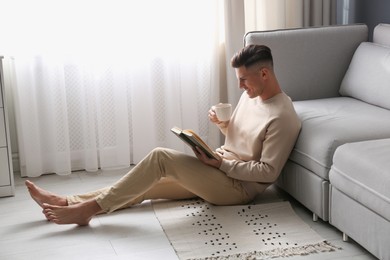 Photo of Man with cup of hot drink reading book at home. Floor heating concept