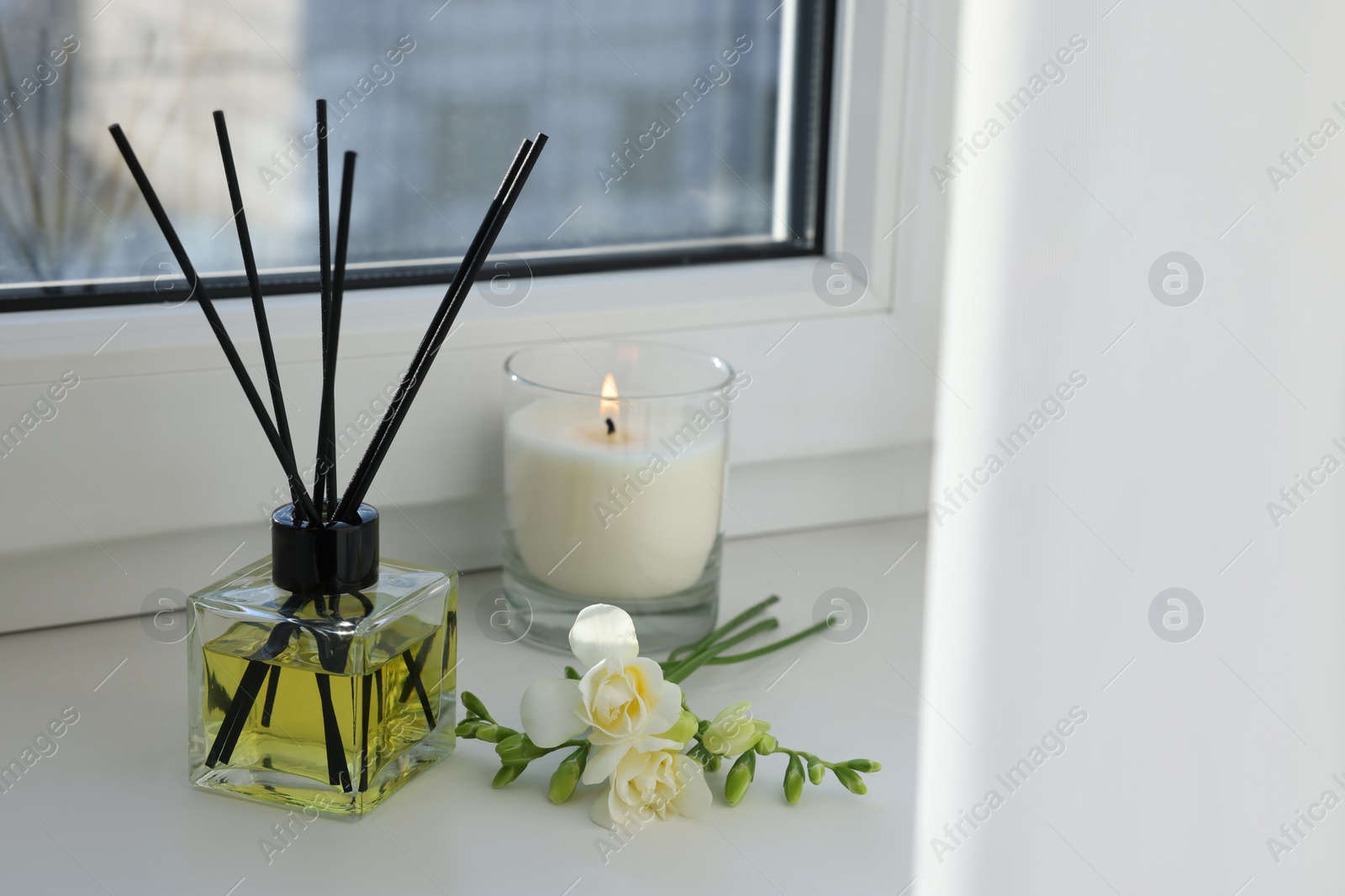 Photo of Aromatic reed air freshener, freesia flowers and burning candle on windowsill