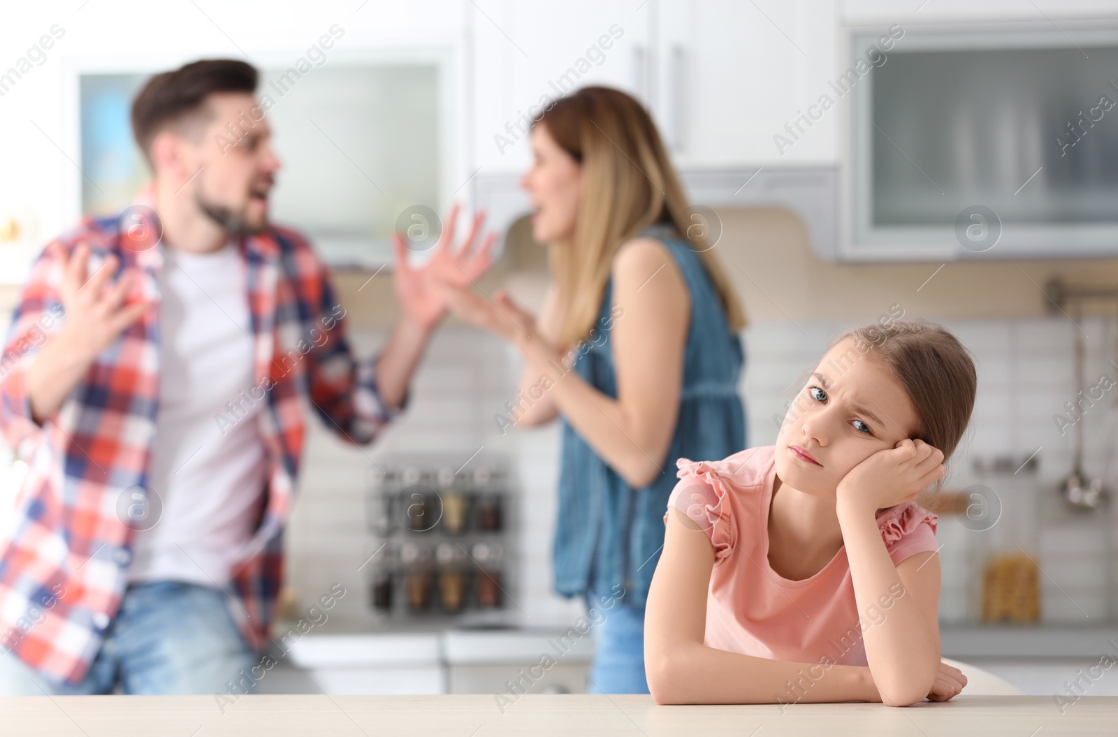 Photo of Little unhappy girl sitting at table while parents arguing on kitchen