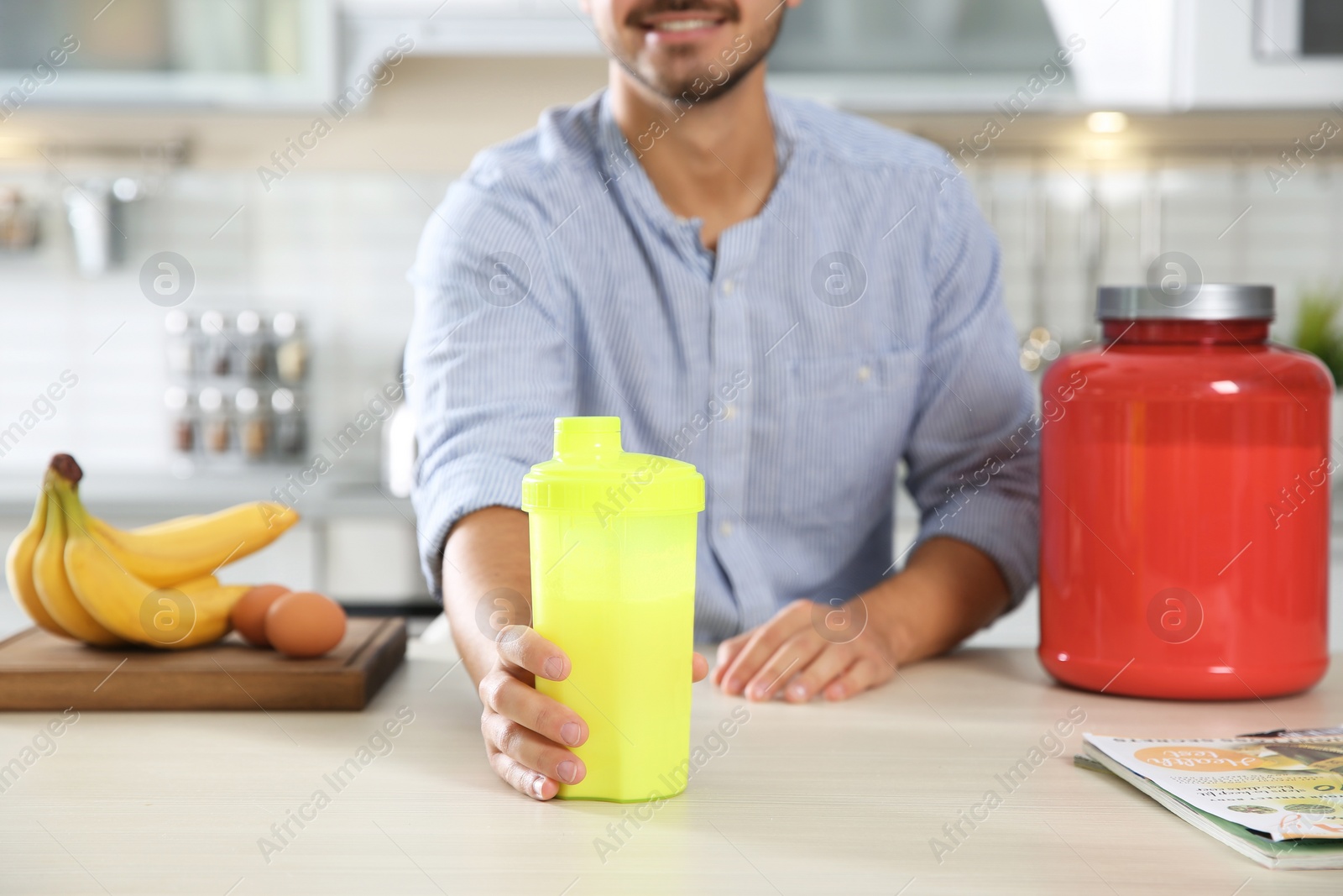 Photo of Young man holding bottle of protein shake at table with ingredients in kitchen