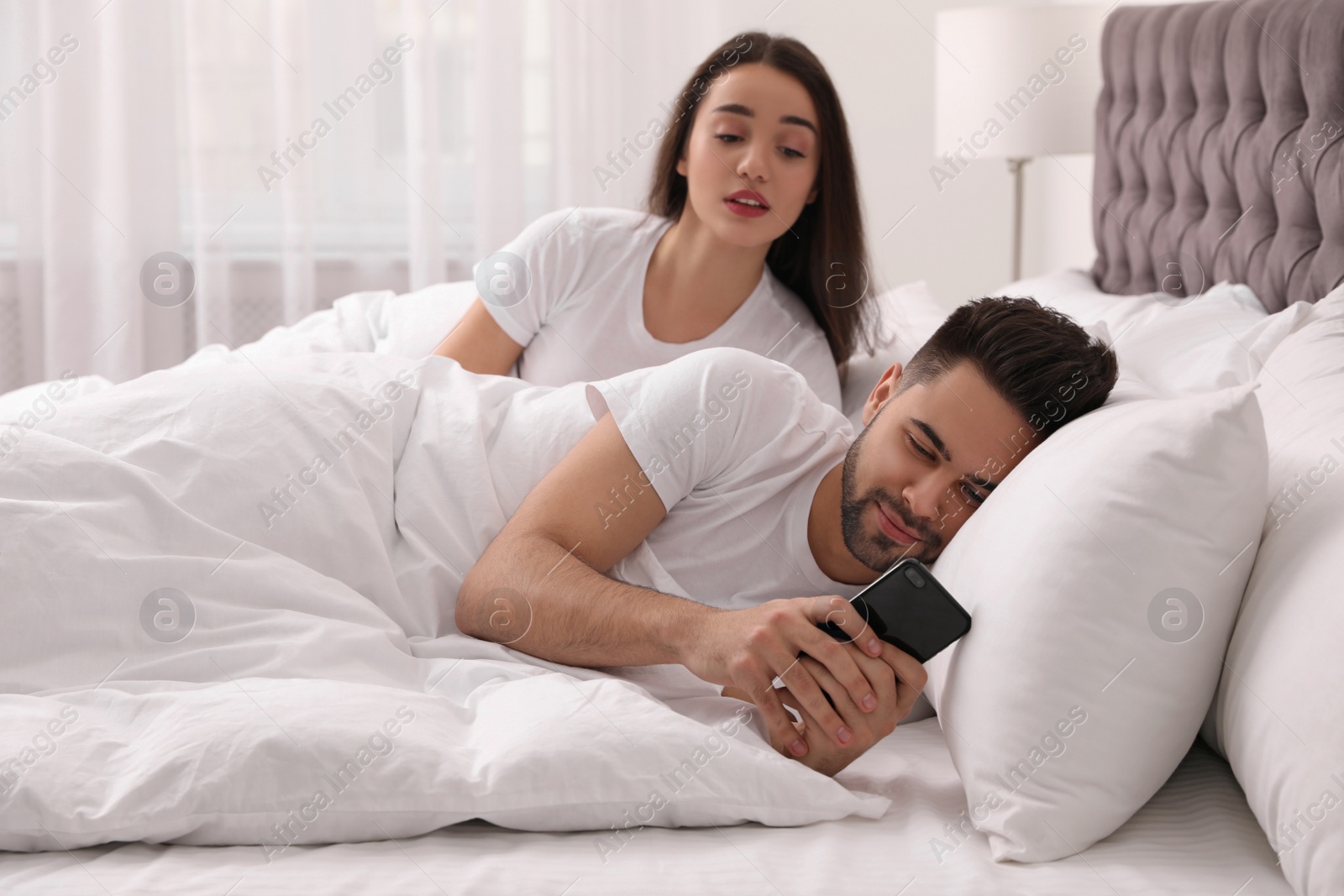 Photo of Distrustful young woman peering into boyfriend's smartphone in bed at home