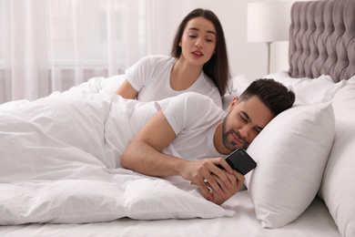 Photo of Distrustful young woman peering into boyfriend's smartphone in bed at home