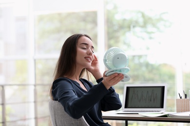 Photo of Young woman enjoying air flow from portable fan at workplace. Summer heat