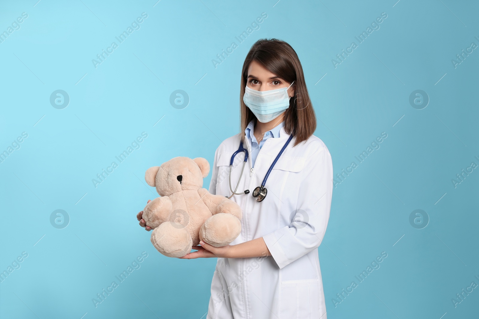 Photo of Pediatrician in protective mask with teddy bear on light blue background