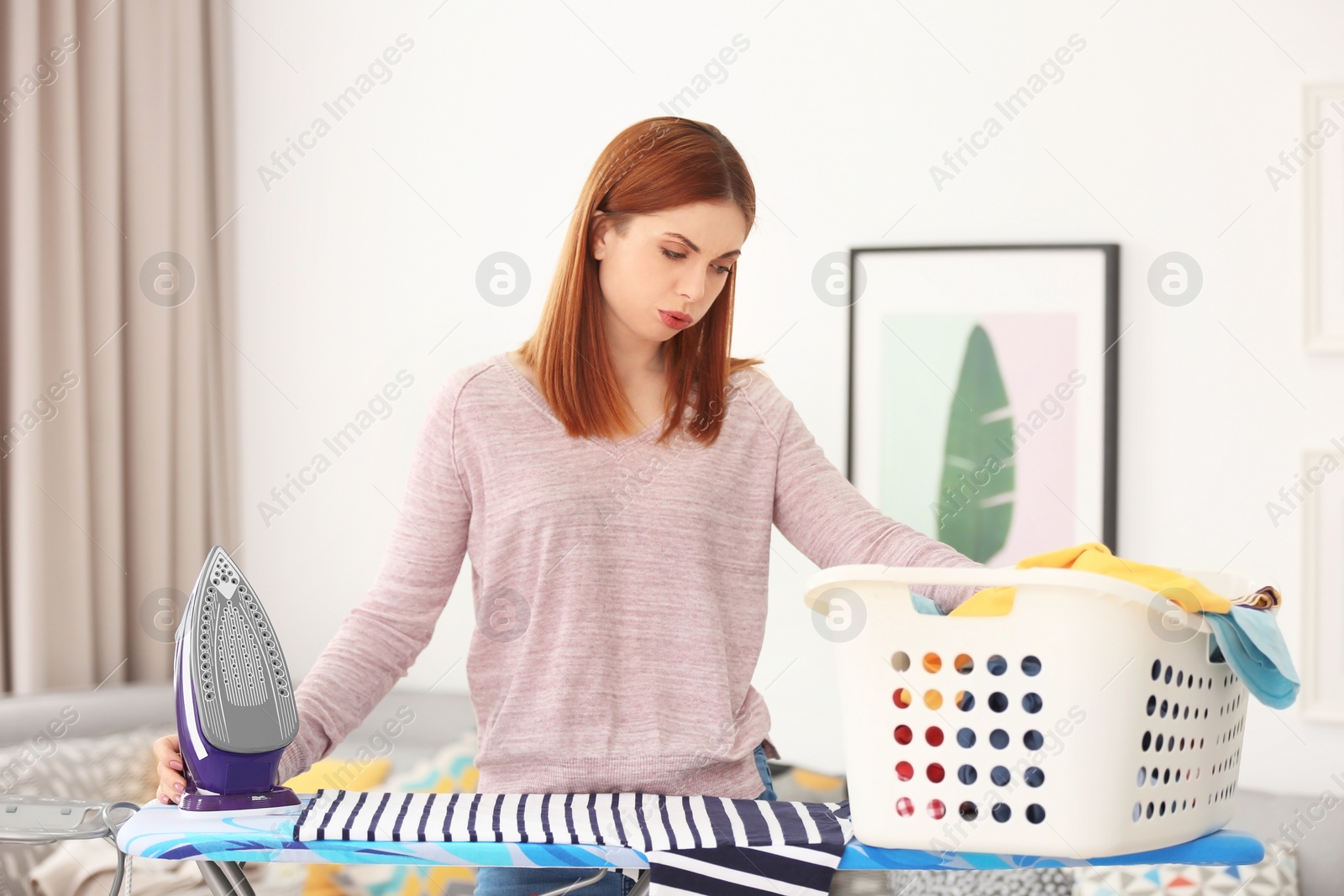 Photo of Tired housewife with basket full of clothes prepared for ironing at home