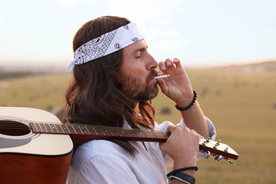 Stylish hippie man with guitar smoking joint in field