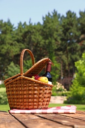 Picnic basket with fruits, bottle of wine and checkered blanket on wooden table in garden