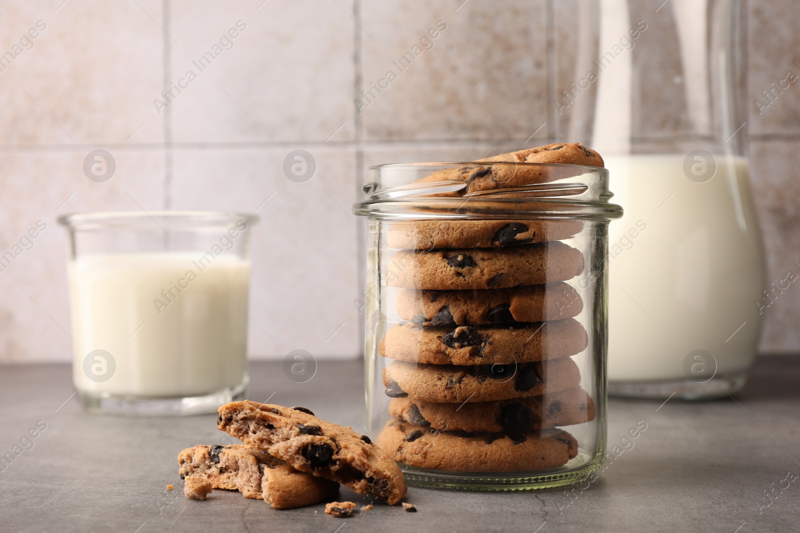 Photo of Glass jar with delicious chocolate chip cookies and milk on grey table