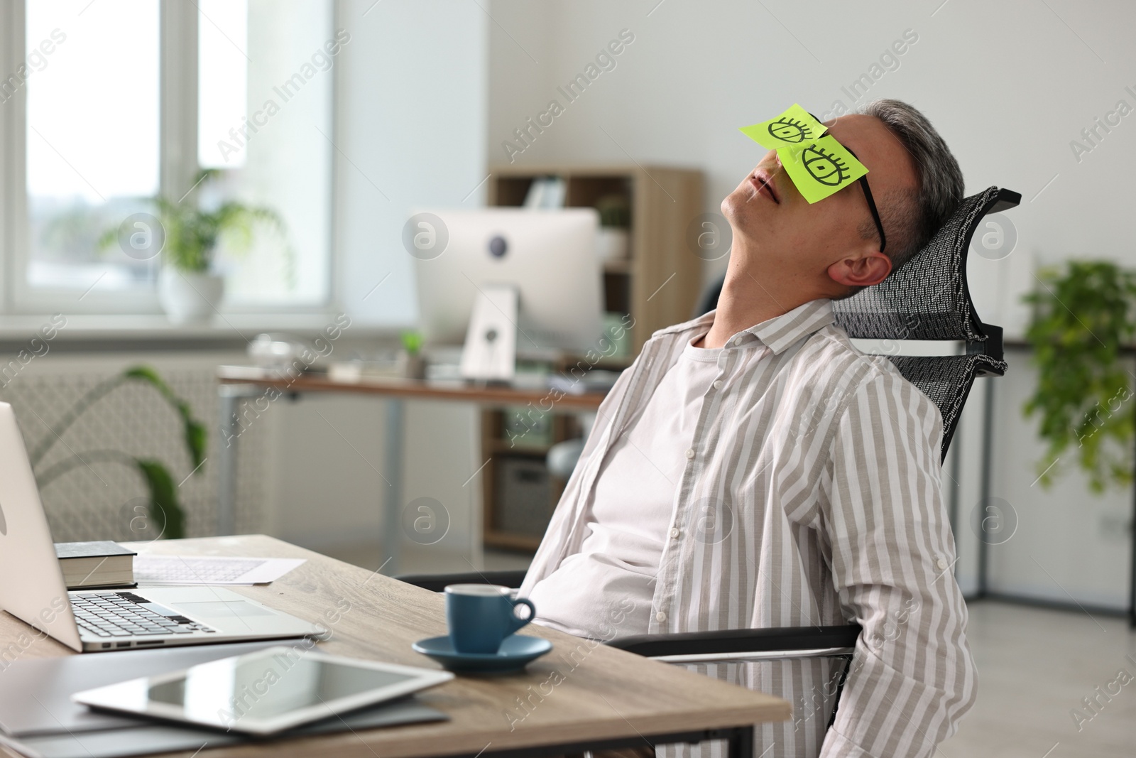 Photo of Man with fake eyes painted on sticky notes snoozing at workplace in office