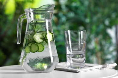 Photo of Refreshing cucumber water with rosemary in jug and glasses on white table against blurred green background, closeup