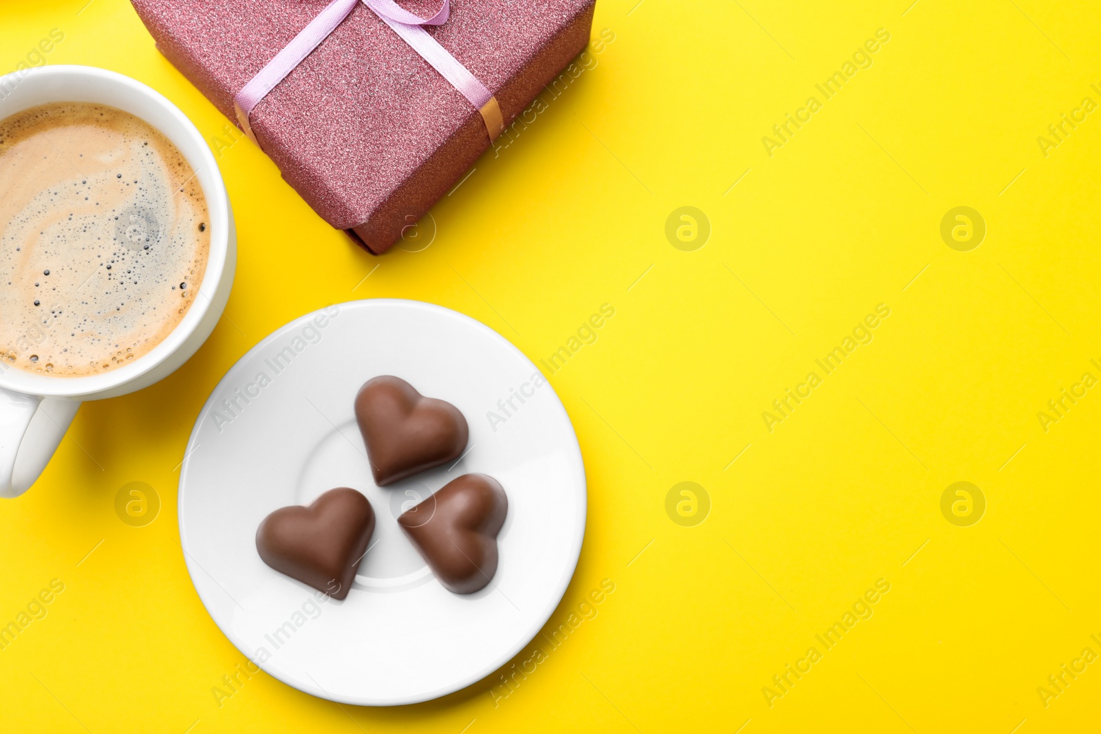 Photo of Cup of coffee, chocolate candies and gift box on yellow background, flat lay with space for text. Valentine's day breakfast